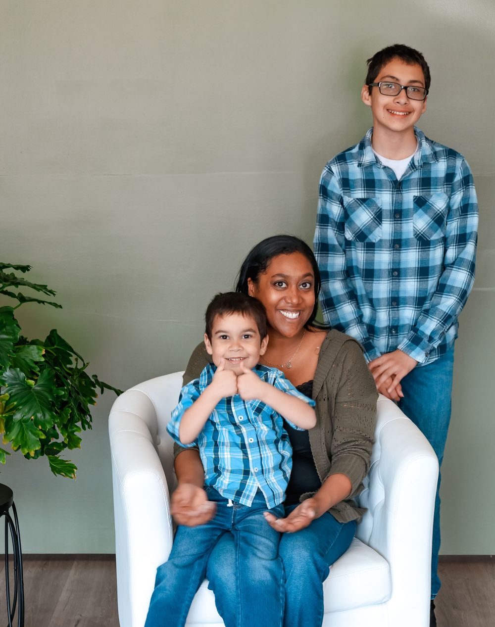 A family of three poses together in a white armchair with a green plant displayed beside them.