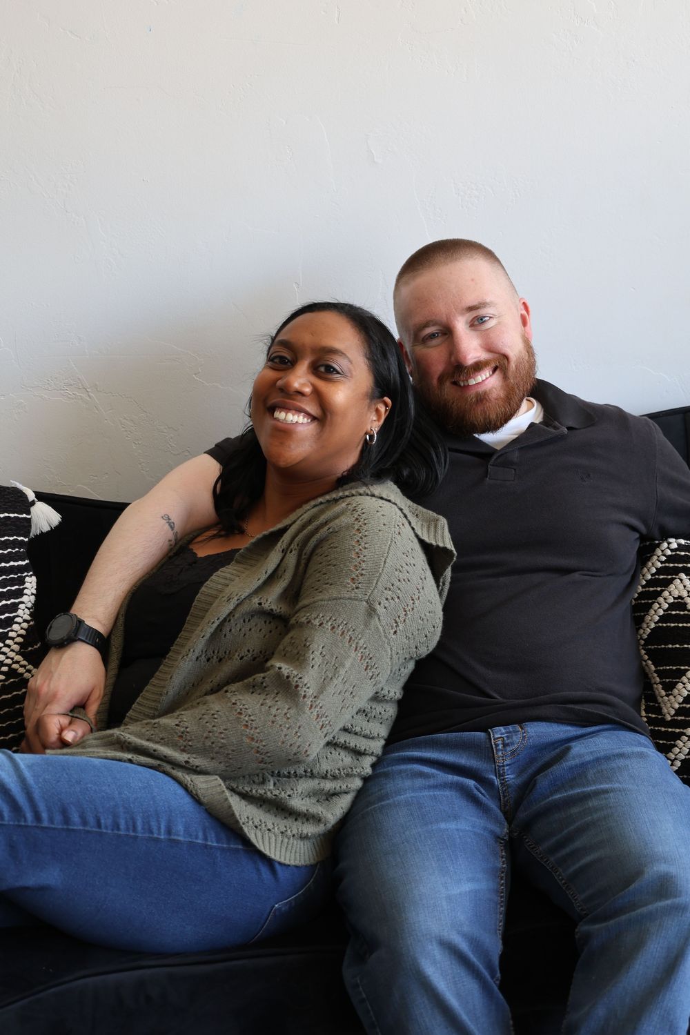 A couple relaxes together on a couch, wearing casual outfits of jeans and sweaters.