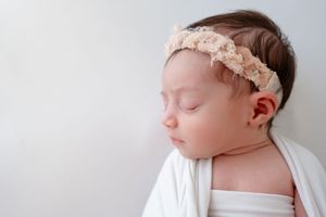Peaceful sleeping newborn wrapped in white cloth wearing a delicate lace headband against light background.