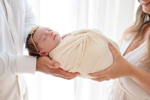 A newborn baby wrapped in a cream knit blanket is cradled gently by hands against a white background.