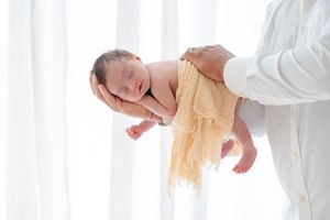 A sleeping newborn baby is gently cradled in someone's arms with soft yellow fabric against a bright window background.