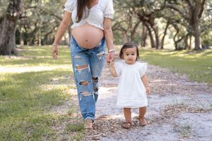 A small child in a white dress holds hands with their pregnant mother wearing ripped jeans on a sunlit path outdoors.