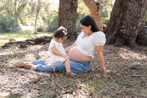 A heartwarming maternity photo showing two people sitting together under a shady tree in dappled sunlight.