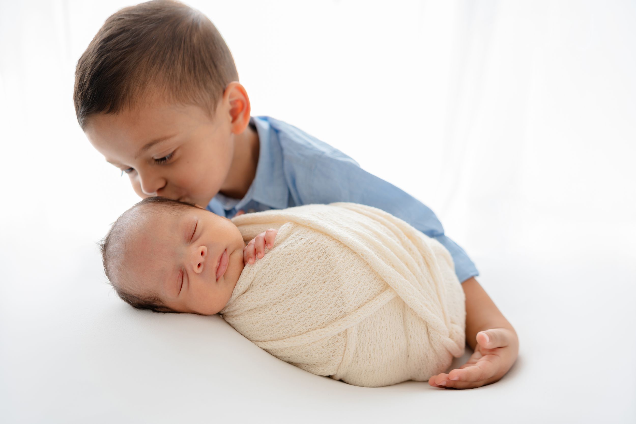 A young child gently embraces a swaddled baby lying on a white background.