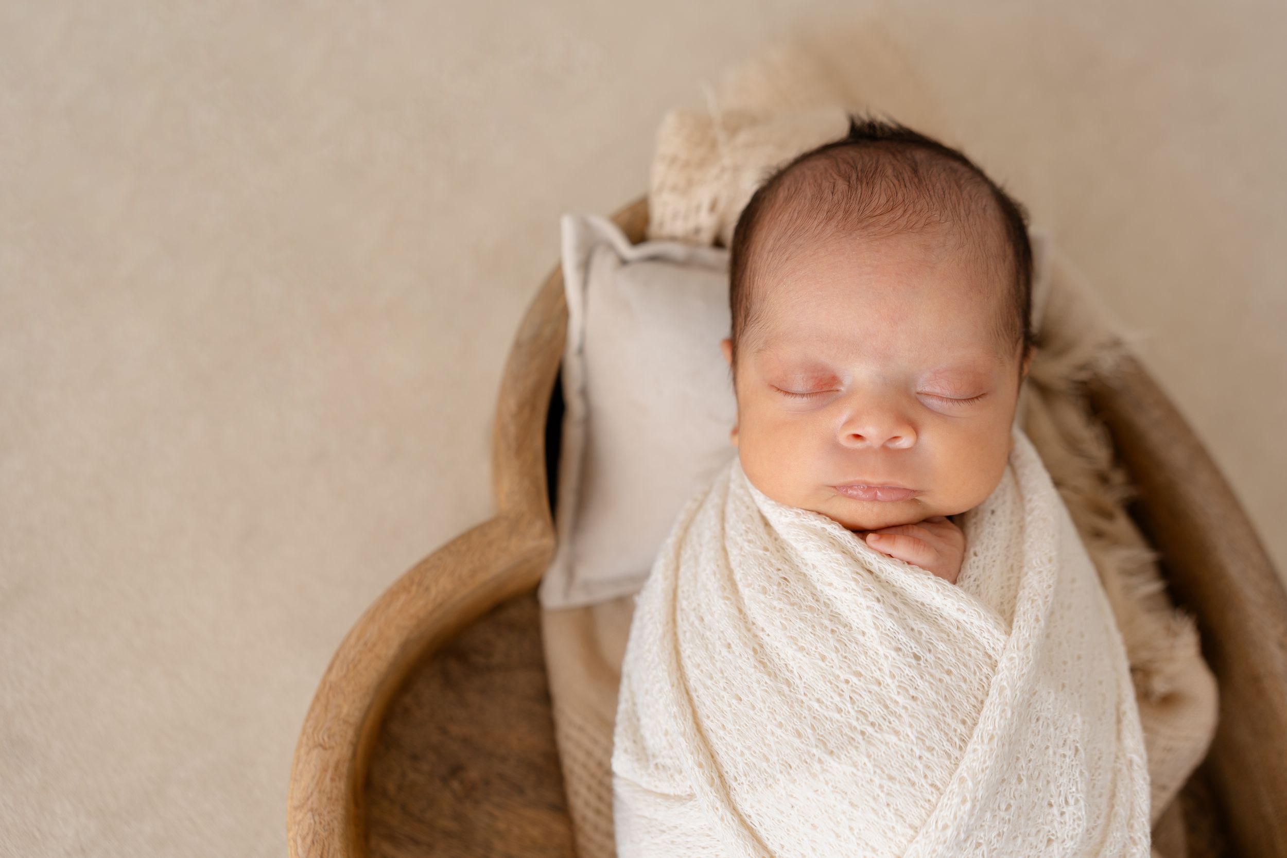 Peacefully sleeping newborn baby swaddled in soft white blanket lying in wooden bowl against neutral backdrop.