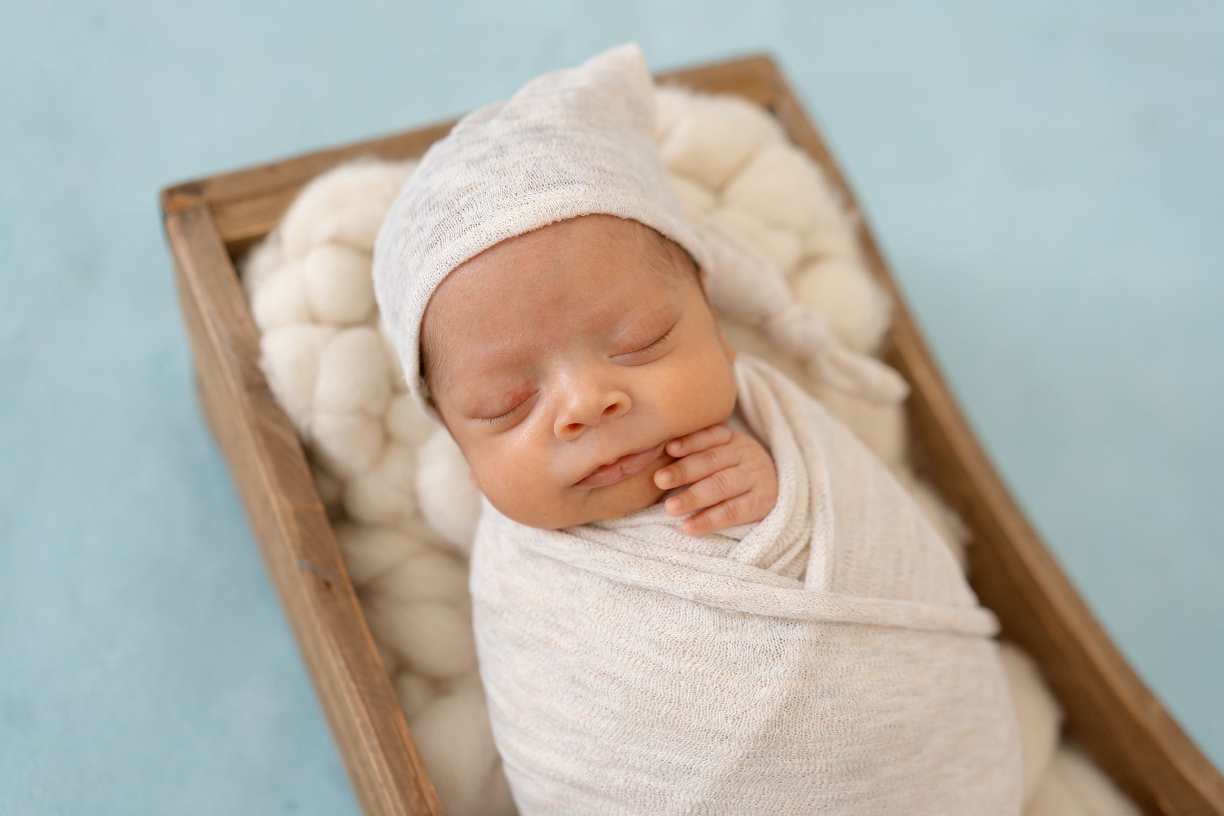 A newborn baby sleeps peacefully in a wooden box while swaddled in soft white fabric with a matching knit hat.