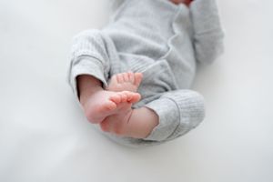 Close up of tiny baby feet with soft gray fabric sleeper pajamas on white bedding.