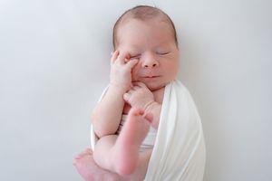 Sleeping newborn wrapped in white cloth rests with hand on face against soft gray background.