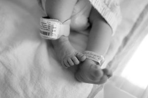 Close-up black and white photo of a newborn baby's tiny feet with hospital identification bracelet on white sheets.