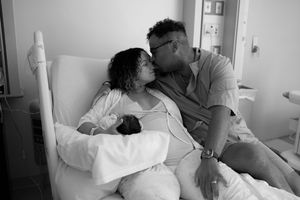 A couple shares an intimate moment in a hospital room while holding their newborn baby in black and white photography.