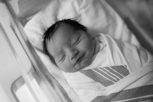 A sleeping newborn baby swaddled in white and striped blankets lies peacefully in black and white photograph.