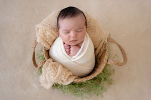 Sleeping newborn wrapped in white swaddle rests peacefully in beige woven basket with green moss accents.