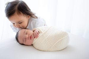 A sweet moment between a child and a sleeping newborn wrapped in a cream blanket on a bright white background.