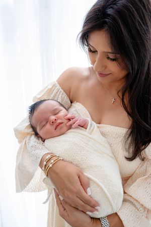 A sleeping newborn wrapped in cream white swaddle cloth rests peacefully in gentle arms wearing a cozy white sweater.