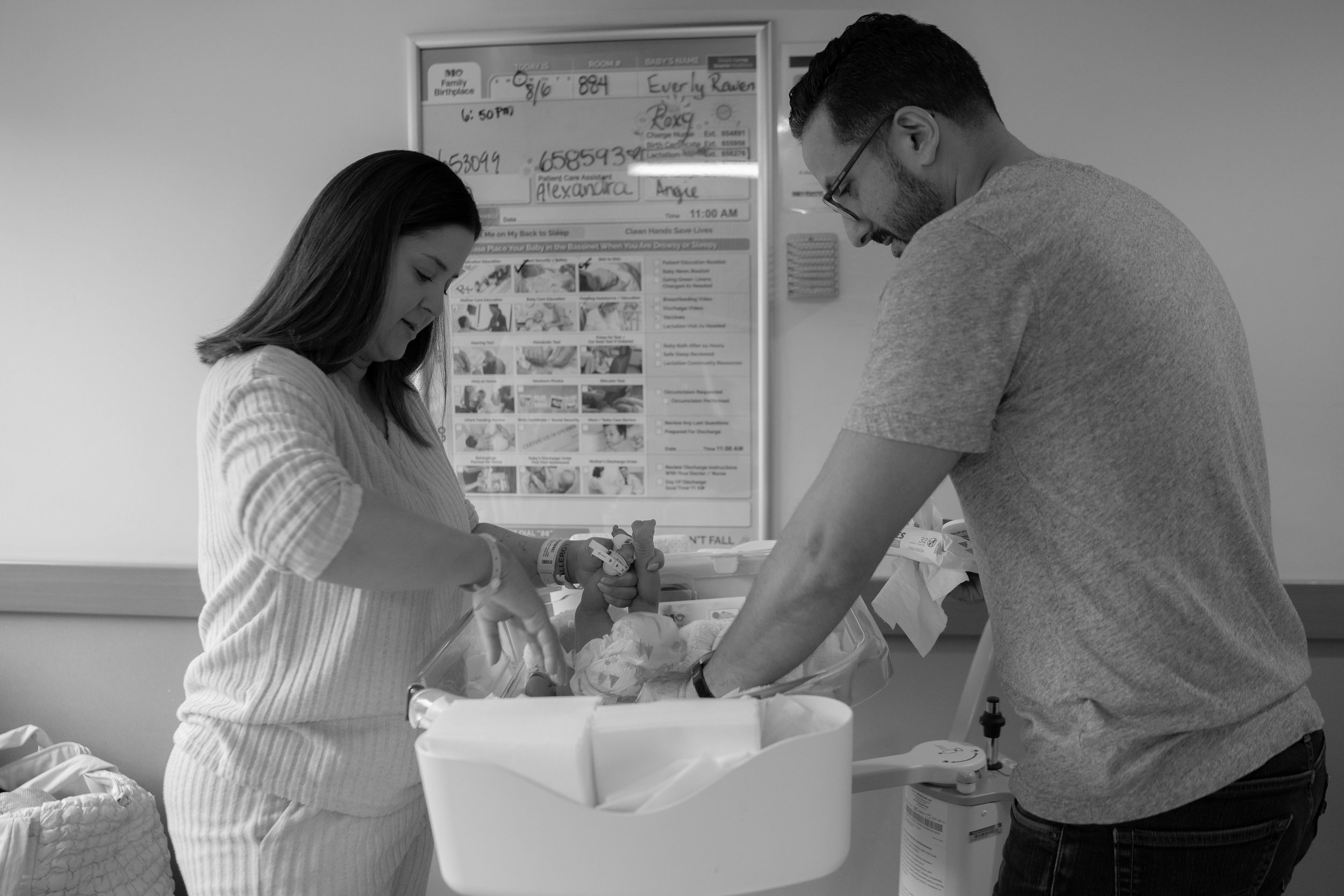 A black and white hospital scene shows two people looking down at a newborn baby in a bassinet.