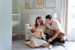 New parents sit on nursery floor with their newborn baby and dog in a sunlit room with minimal wall art above.