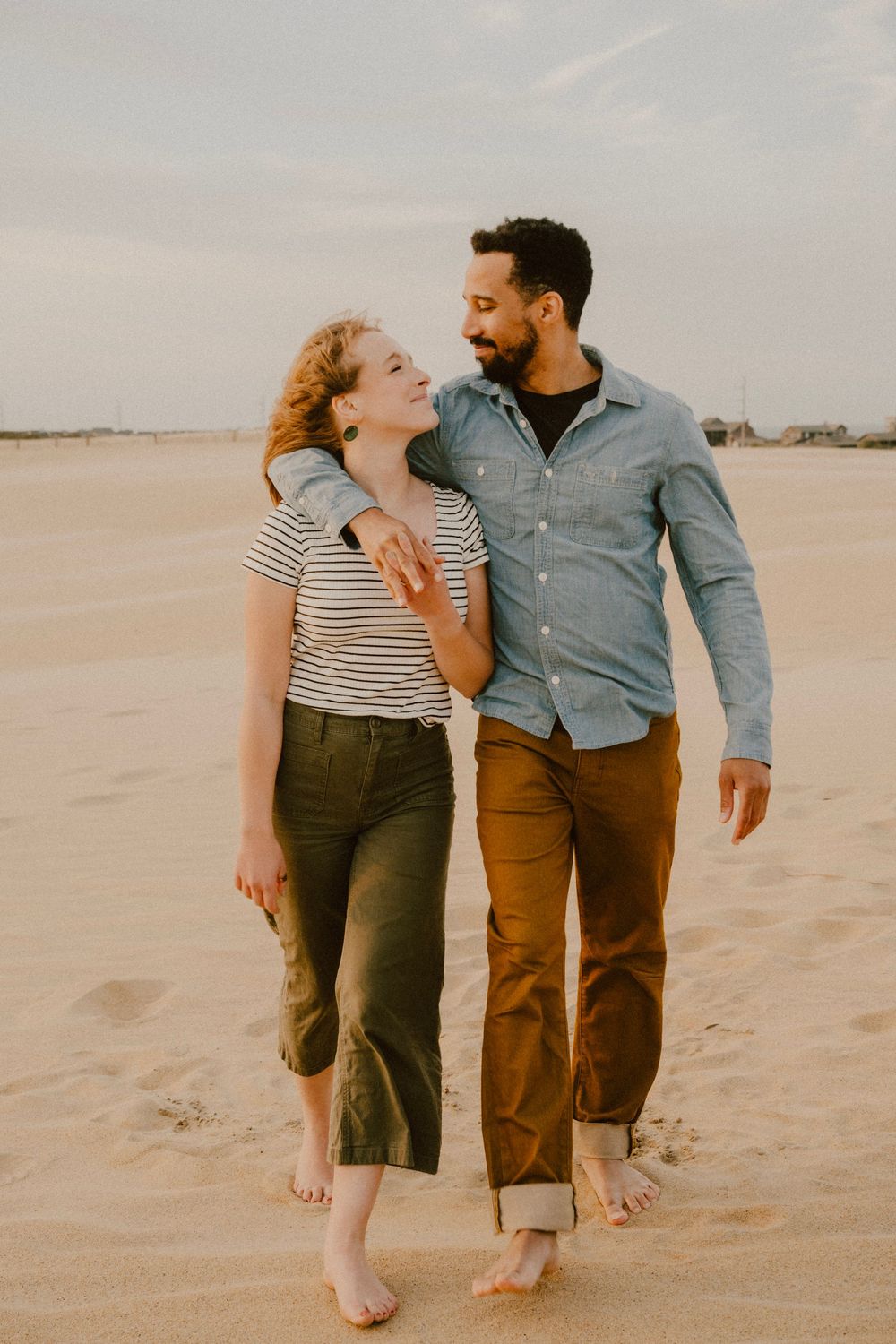 Couple in casual attire walking barefoot together on sandy beach.