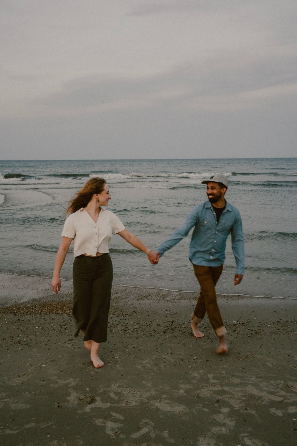 Couple holding hands while walking barefoot along ocean shoreline.