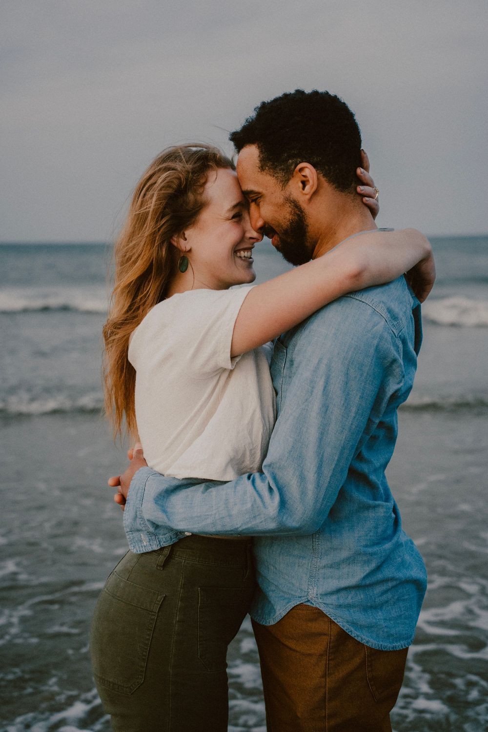 Romantic couple embracing on a beach at sunset, wearing coordinating casual outfits.
