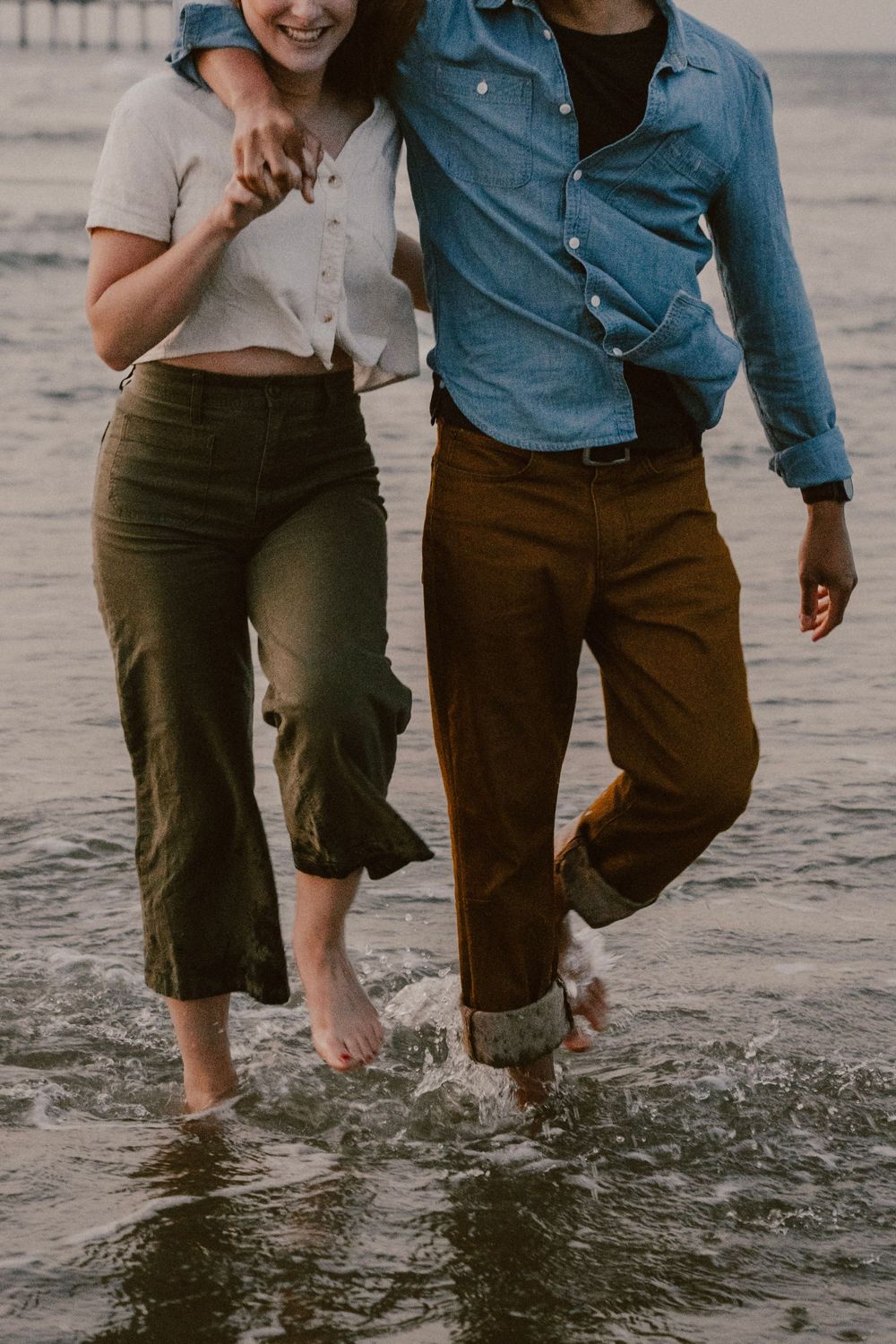 Couple splashing through ocean waves, wearing matching button-downs and casual pants on a beach.