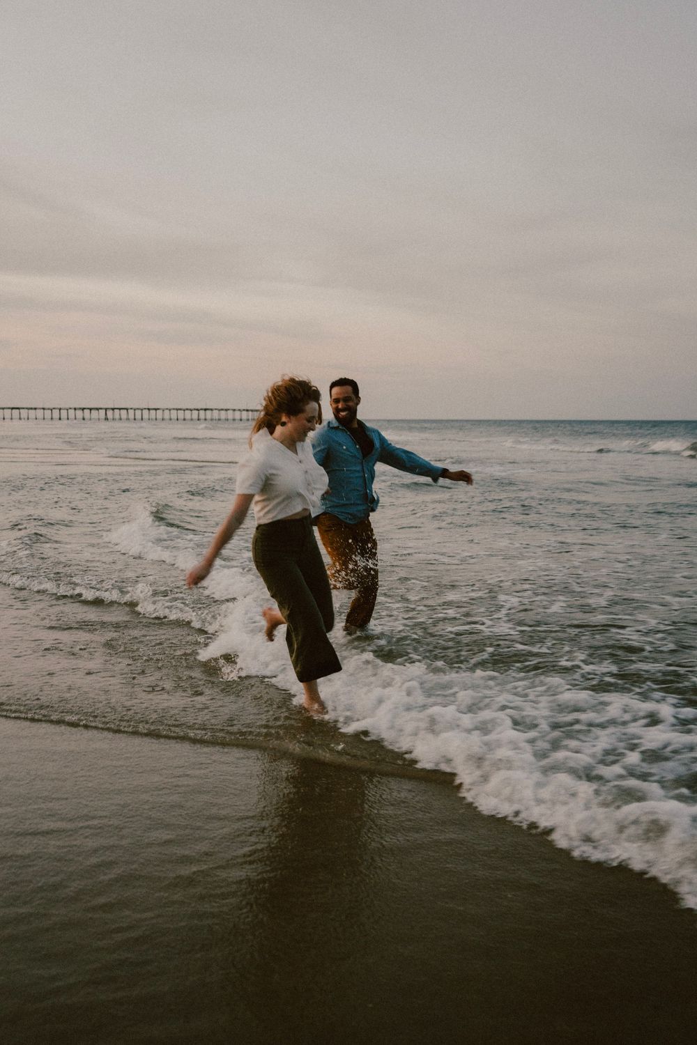 Playful couple running through ocean waves on a beach with a pier in the background.