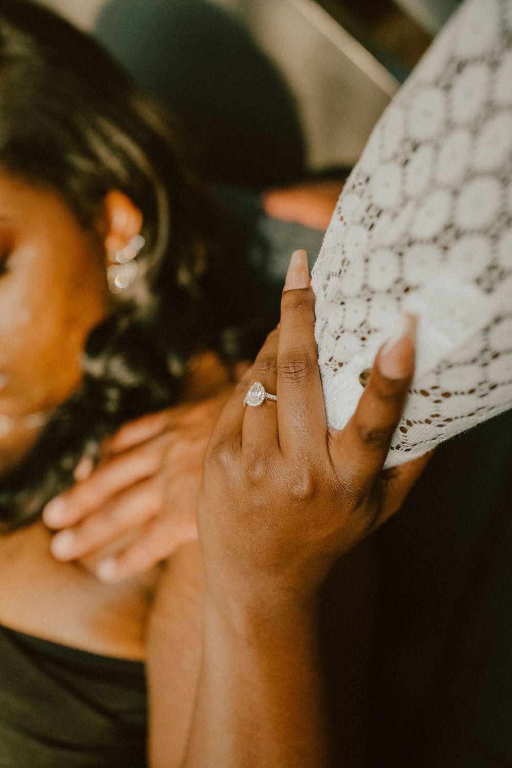 Close-up of a diamond engagement ring on hand against a lace background in warm lighting.