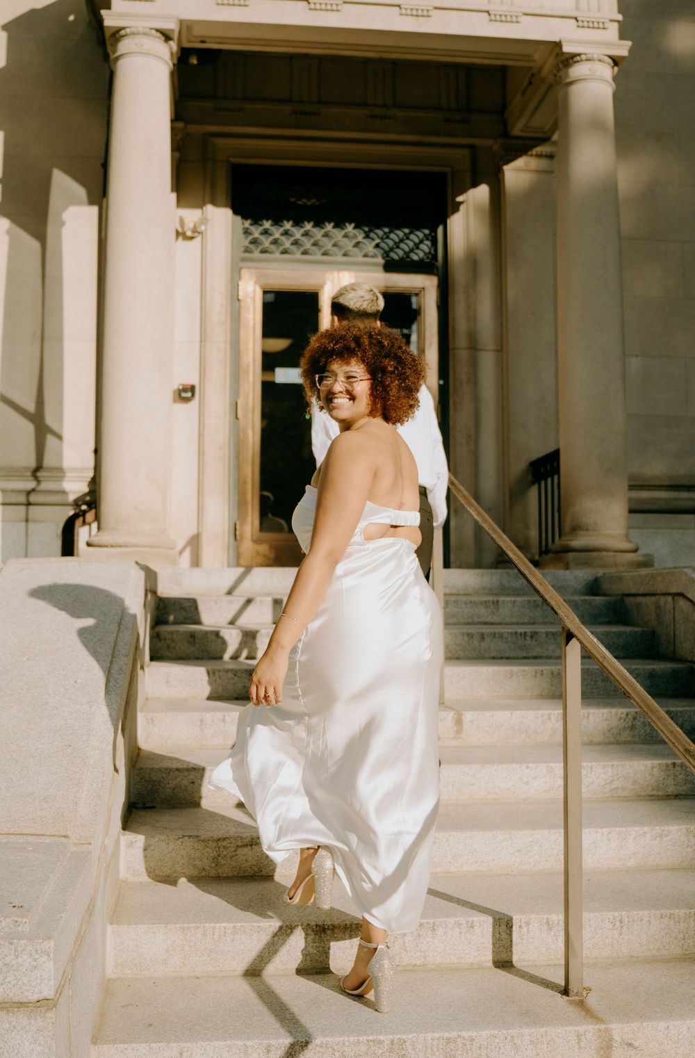 A woman in a flowing white dress poses on grand marble steps of a building entrance.