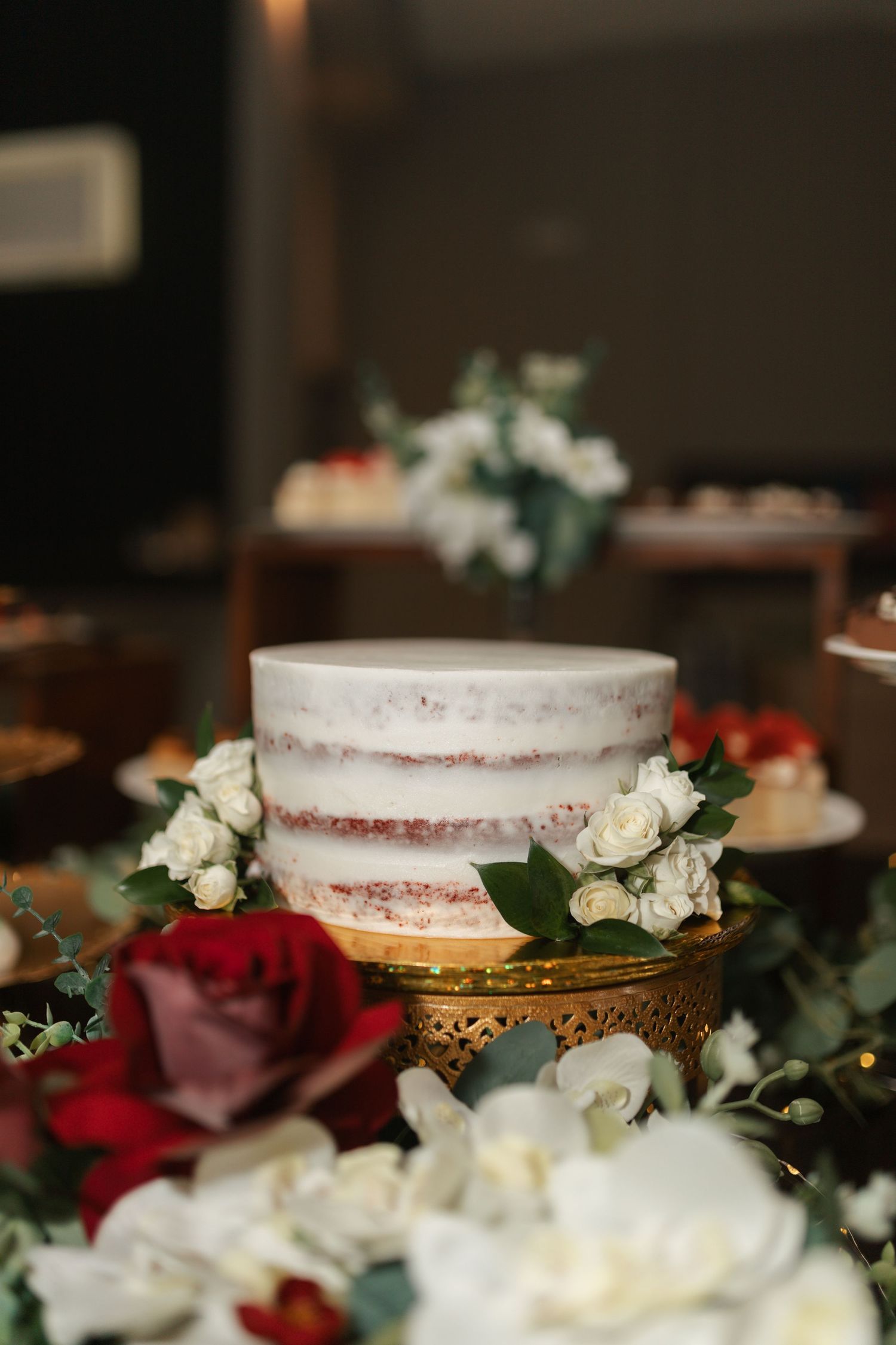 Elegant semi-naked wedding cake decorated with white roses and greenery on gold stand.