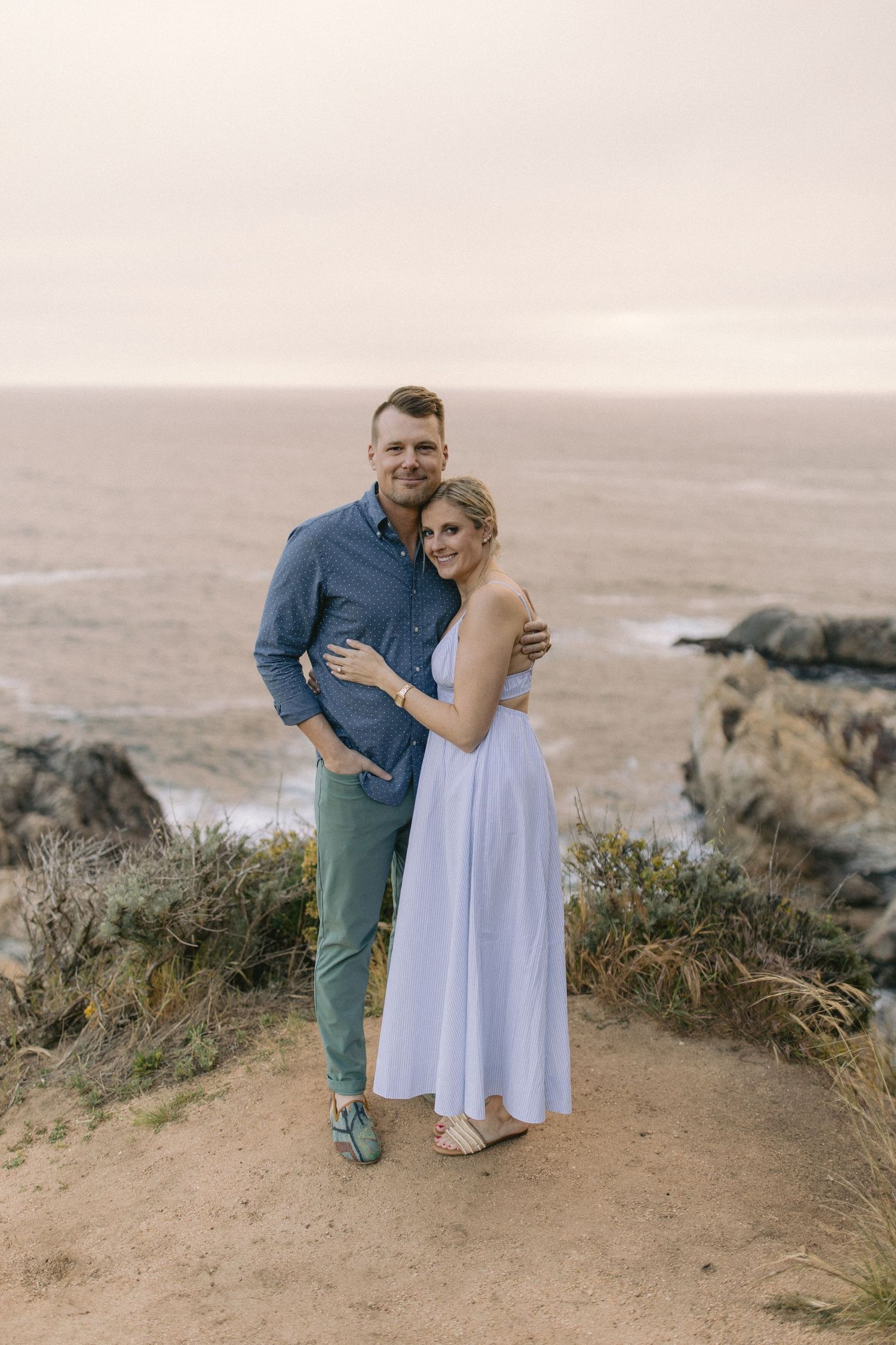 A couple embraces while standing on a coastal cliff overlooking the ocean at sunset in casual formal attire.