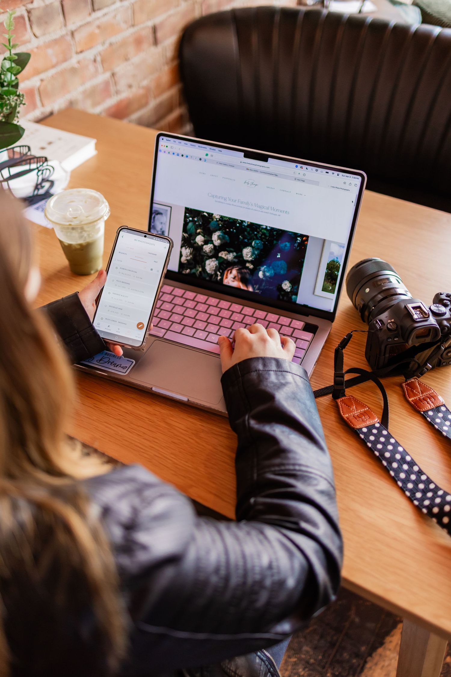 Photographer working on laptop with pink keyboard, camera and iced coffee at wooden desk in cafe.