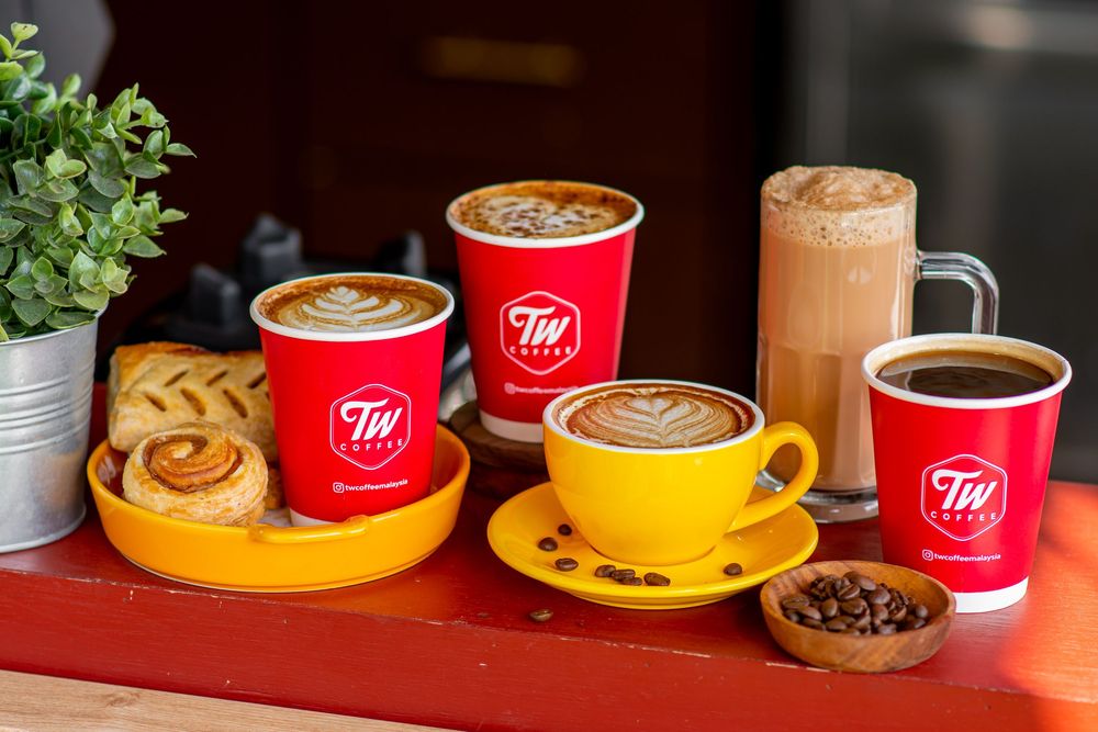 Row of red and yellow coffee cups arranged on saucers with coffee and plants in background.