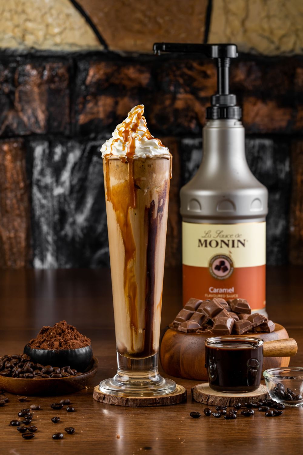 Caramel frappe topped with whipped cream alongside Monin syrup and chocolate pieces.