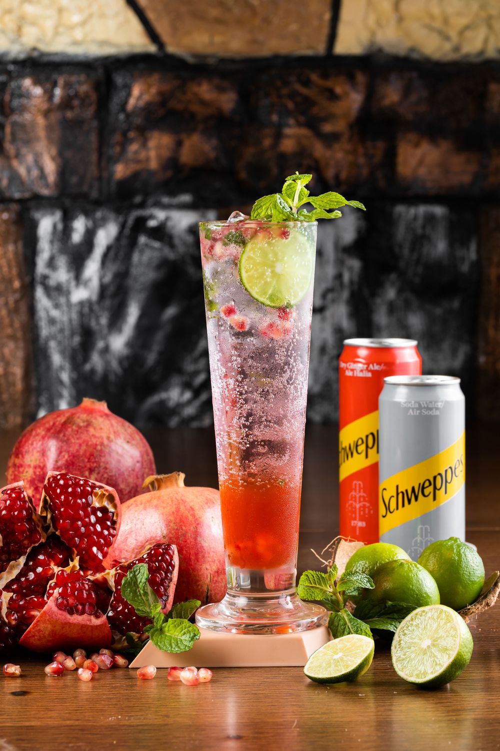 Refreshing pomegranate cocktail with lime garnish and Schweppes cans on wooden table.