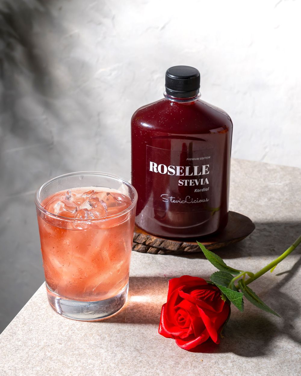 Pink cocktail with ice in a glass next to a dark bottle and red rose on a light surface.