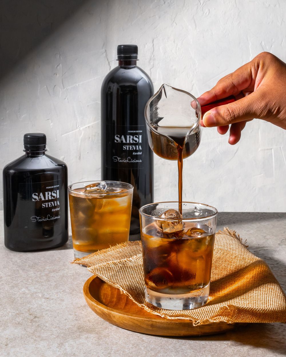 Pouring dark spirits into glasses with ice on a wooden serving board.