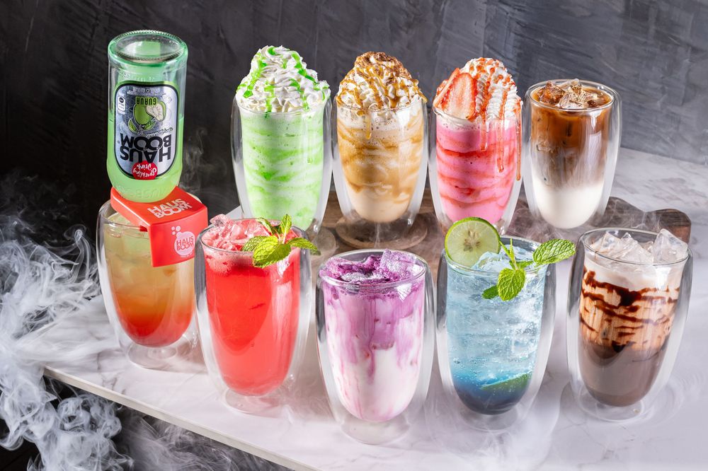Collection of colorful iced beverages with whipped cream toppings on smoky display.