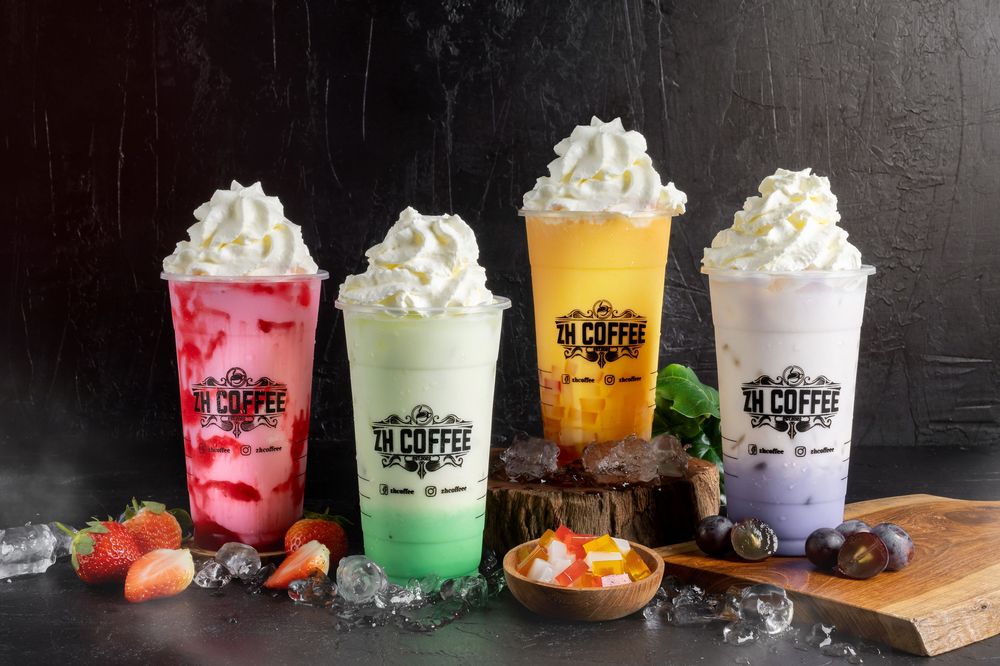 Colorful array of whipped cream topped beverages in clear cups on dark surface.