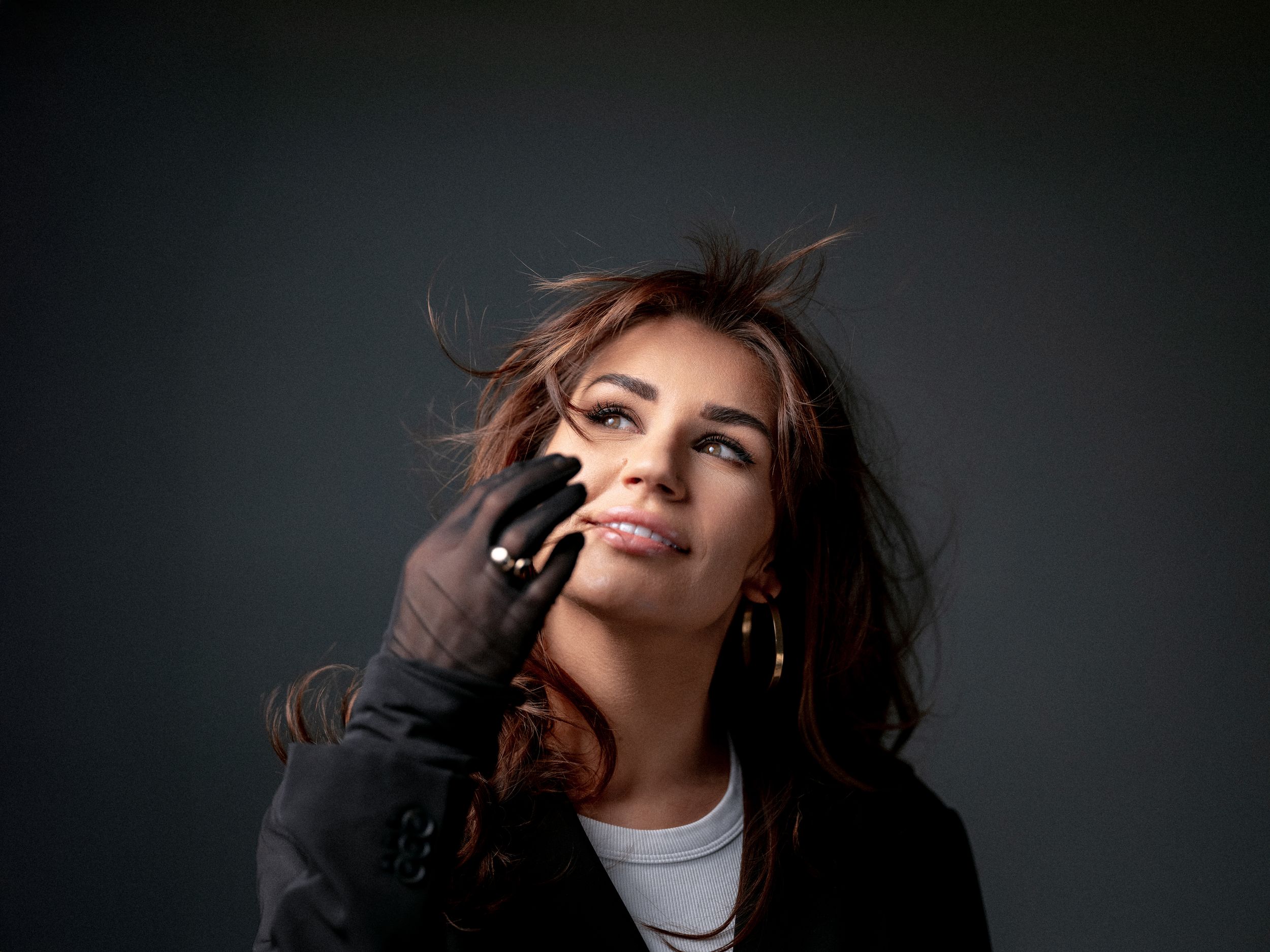 Dramatic studio portrait with flowing dark hair against black background wearing black gloves and white top.