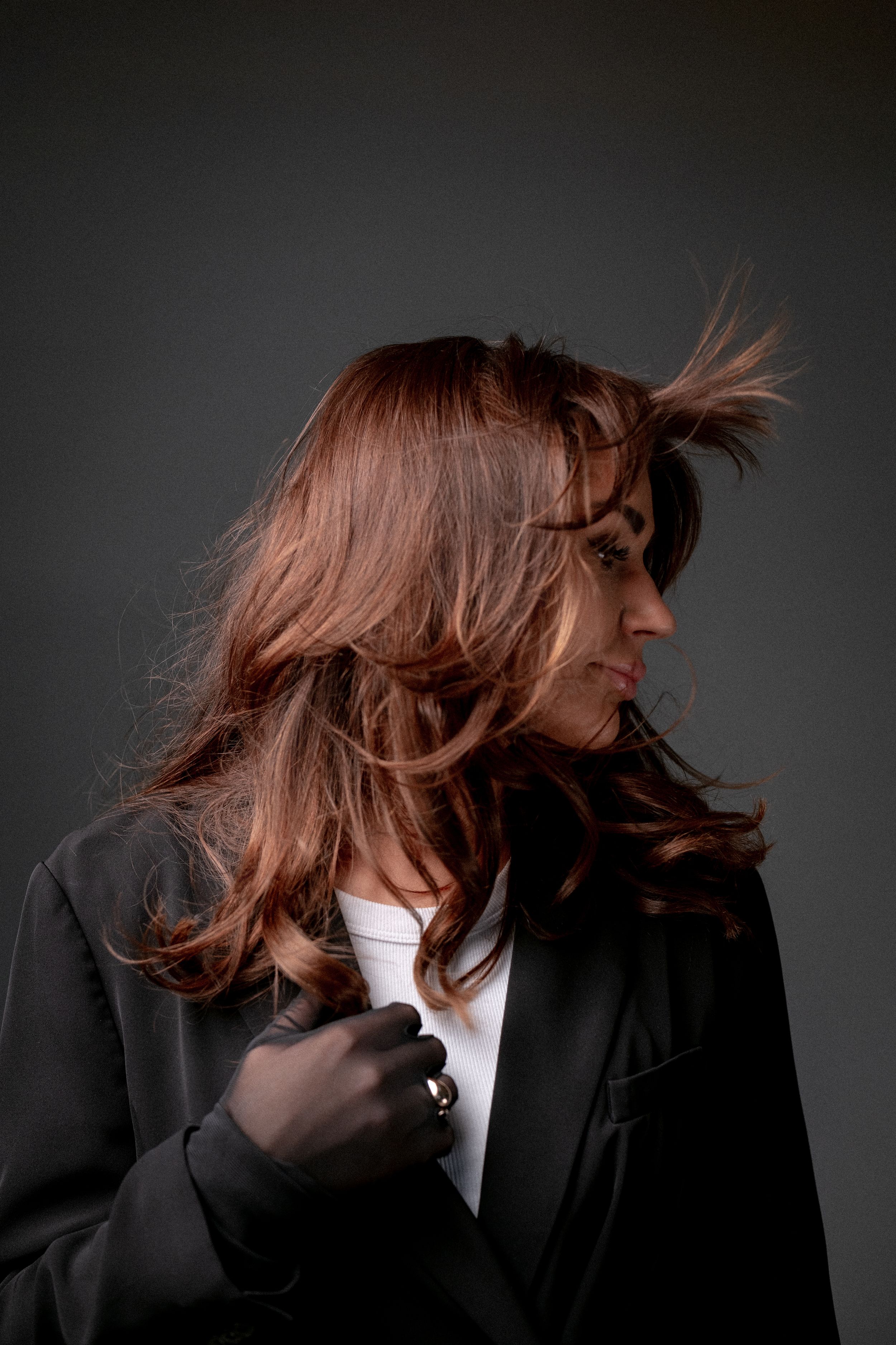 Model posing in dark black blazer with flowing reddish-brown hair against a gray studio background.
