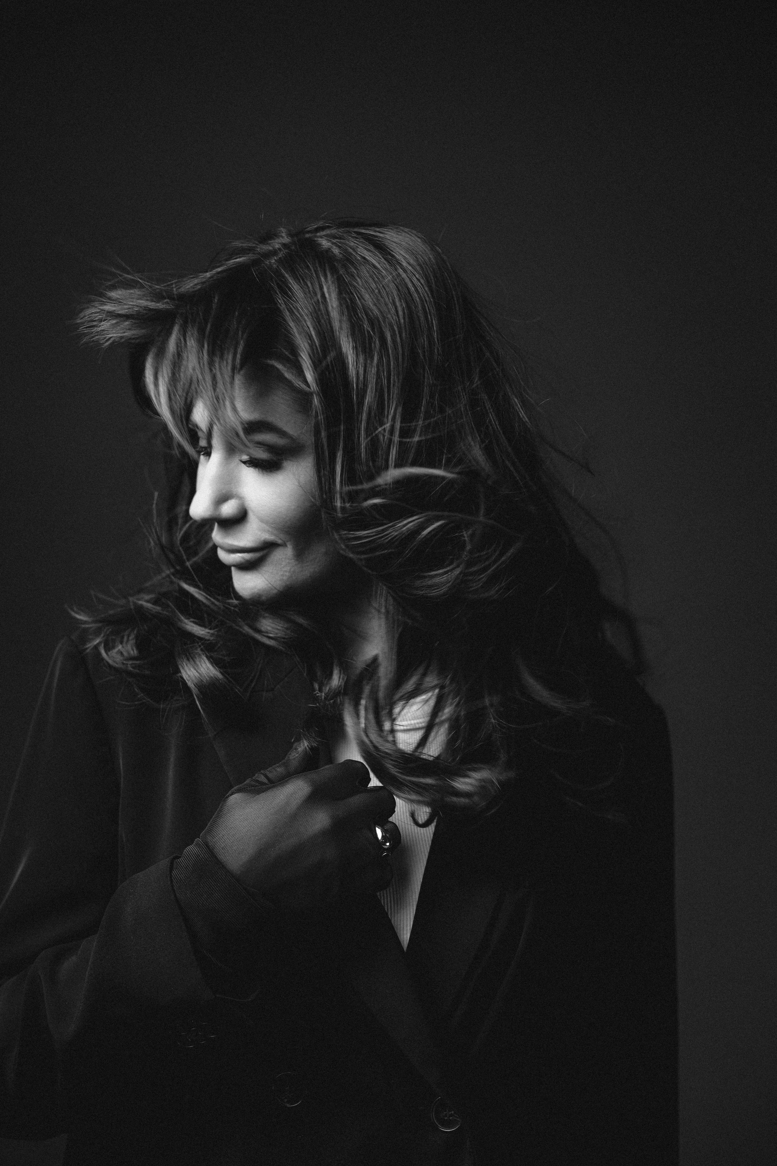 Dramatic black and white portrait of person with windswept hair in dark clothing against black background.