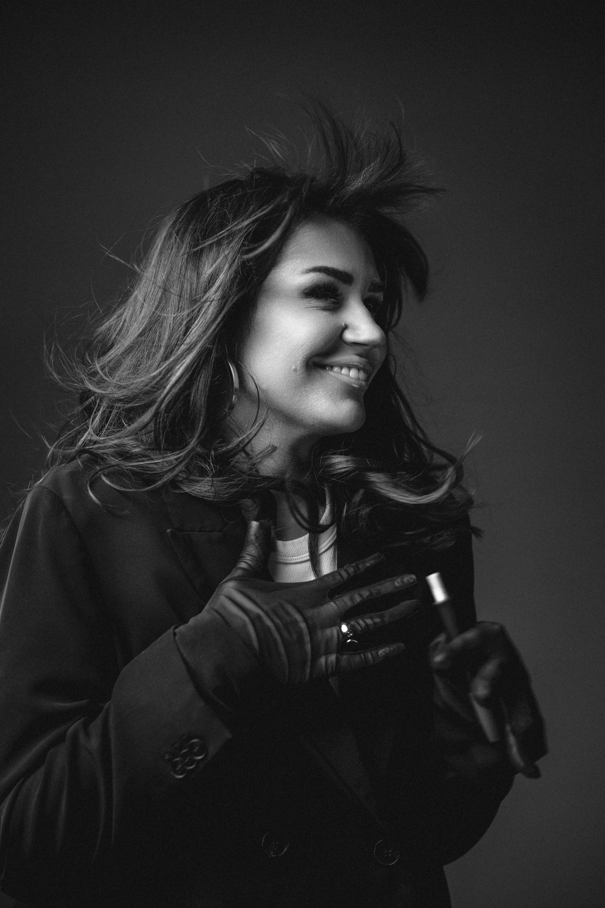 A black and white portrait of a smiling woman with windswept hair wrapped in a dark coat and gloves.