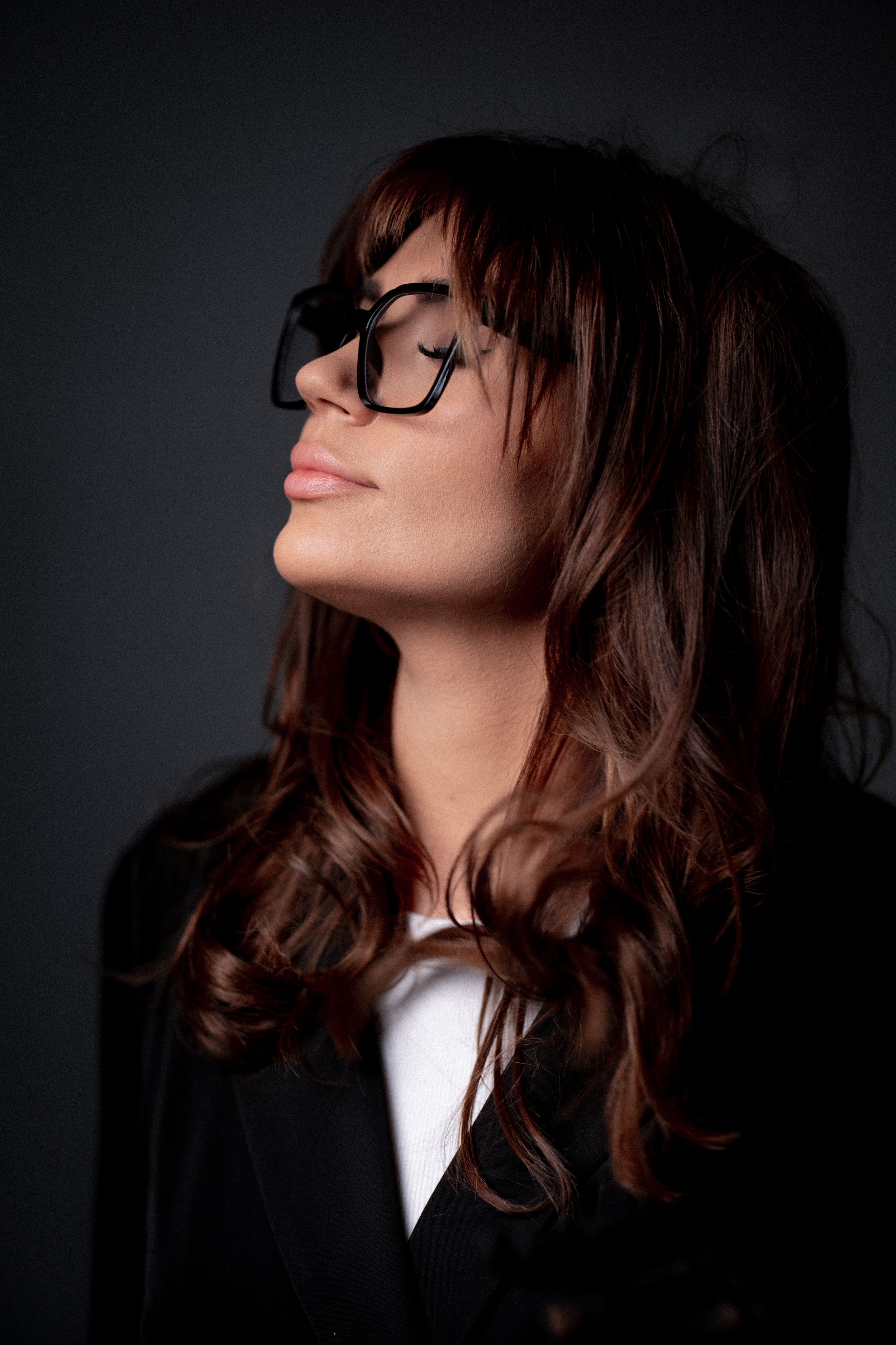 Profile view of a figure with wavy brown hair and glasses tilting head back against dark background.