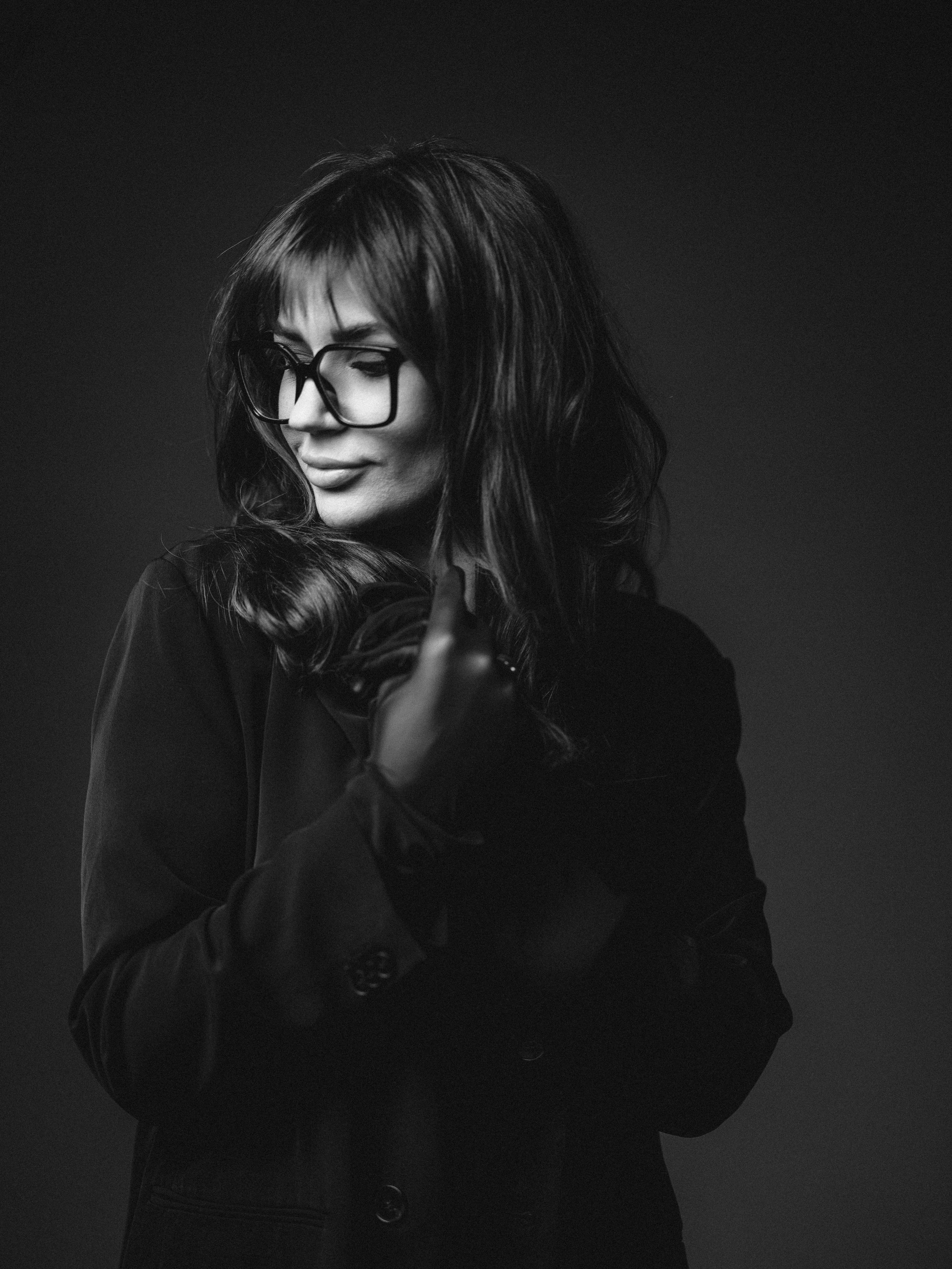 Dramatic black and white portrait of a person with glasses and flowing dark hair wearing a black jacket against dark background.