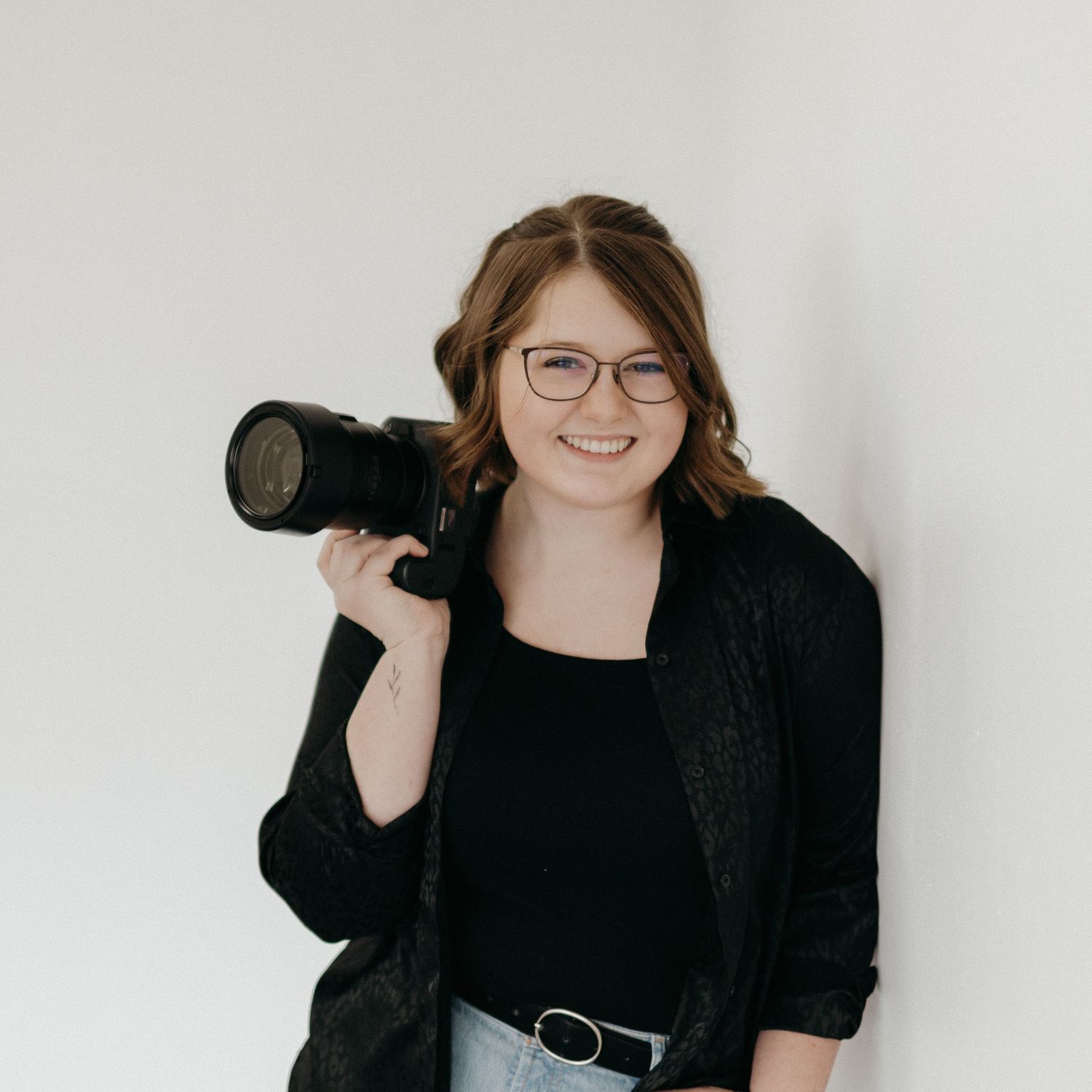 Professional photographer wearing glasses and black attire holds a camera with telephoto lens against white background.