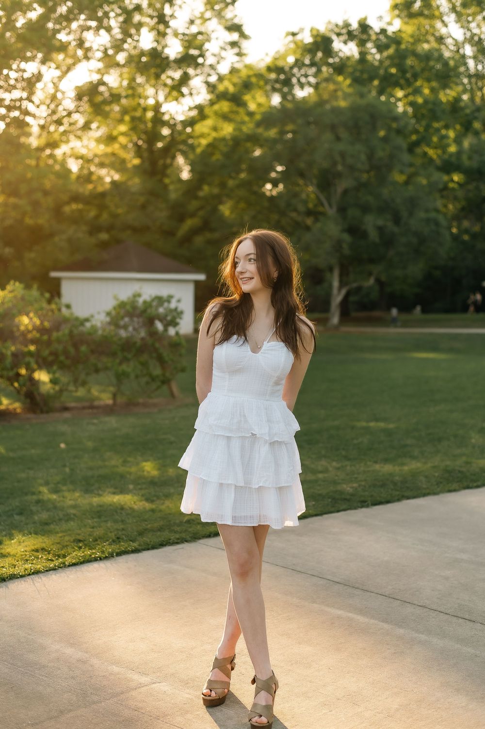 Woman in white dress standing on concrete path during sunset with trees in background.