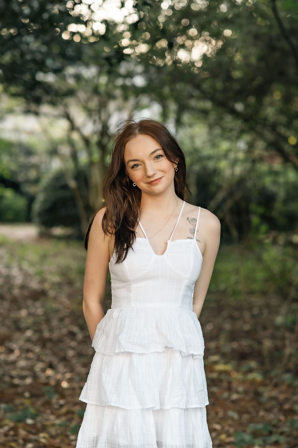 Woman in white summer dress poses in wooded garden setting with soft lighting.