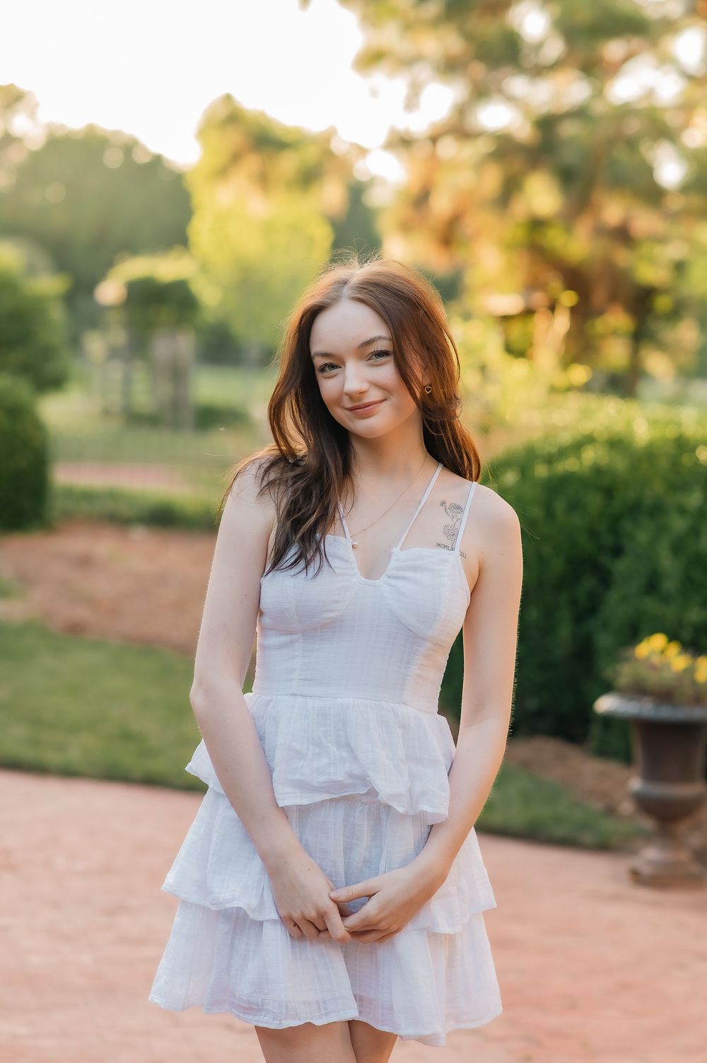 Woman in white sleeveless dress poses in garden with sunset lighting.