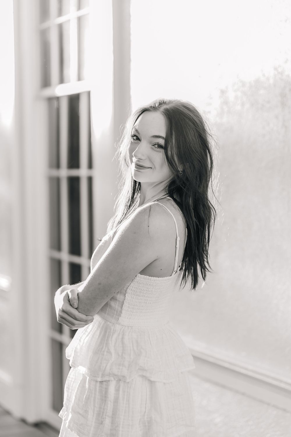 Black and white portrait of woman in white dress standing by window.