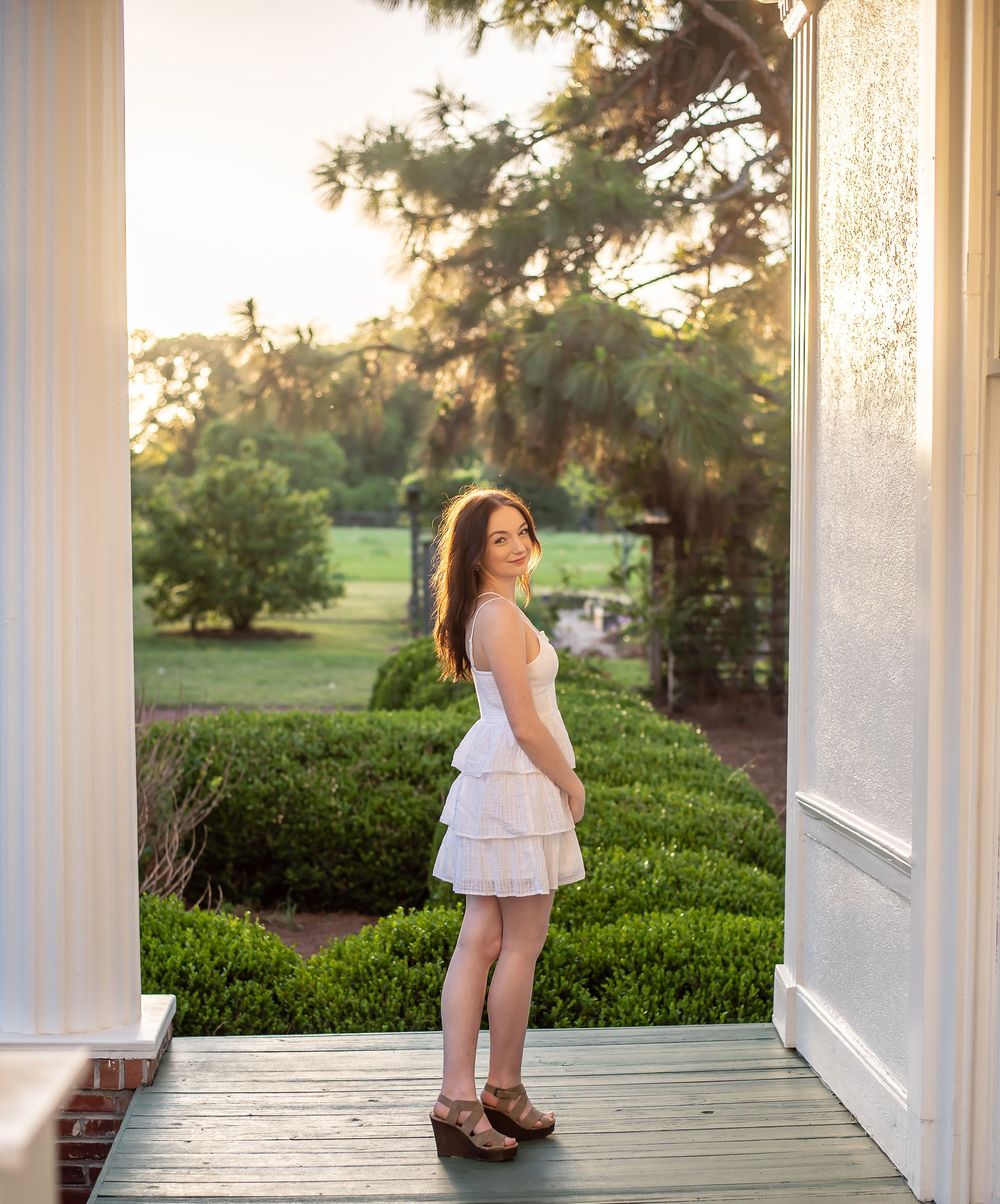 Woman in white dress standing on porch with garden view during sunset.