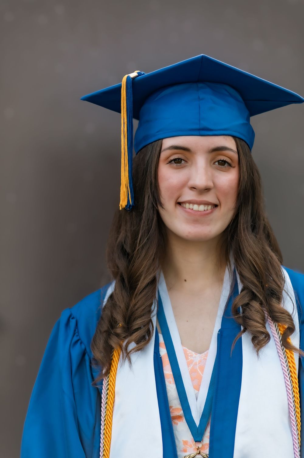 Graduate wearing blue cap and gown with gold tassel against dark background.